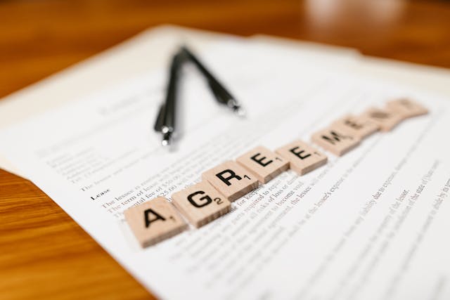 a lease on a wooden table with two pens on top of it and scrabble tiles laid out that spell "agreement"